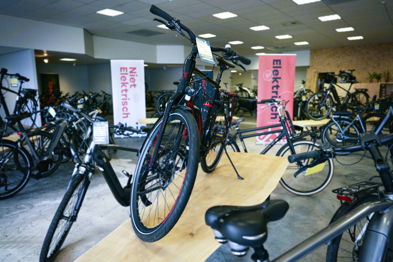 A showroom displaying refurbished ex-lease bicycles, both electric and non-electric, categorized with banners. The restored bikes are showcased on wooden platforms, emphasizing their second life after refurbishment. This initiative, in collaboration with custom work companies, promotes sustainability by extending product lifespan and supporting a circular economy.