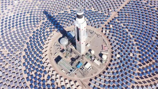 A solar thermal power plant with a central tower surrounded by heliostat mirrors in a radial pattern, set in a desert landscape.