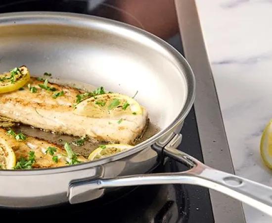 A stainless steel pan with fried fish fillets, garnished with lemon slices and parsley, on an induction cooktop. The pan has a nonstick texture as a PFAS-alternative.